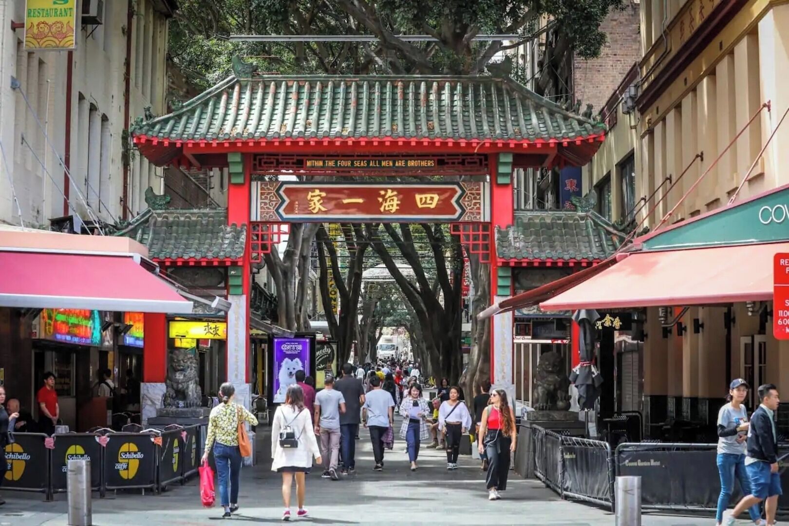 Une porte chinoise traditionnelle, avec un toit rouge et des caractères chinois dorés, qui mène à une rue animée bordée de boutiques et de cafés.