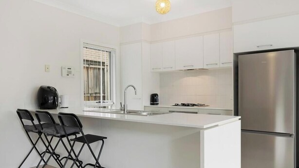 Una cocina moderna con muebles blancos, un refrigerador de acero inoxidable, estufa a gas y una zona de bar con sillas plegables.