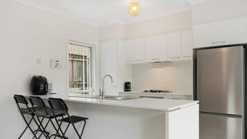 A modern kitchen with white cabinetry, a stainless steel refrigerator, a gas stove, and a bar area with folding chairs.