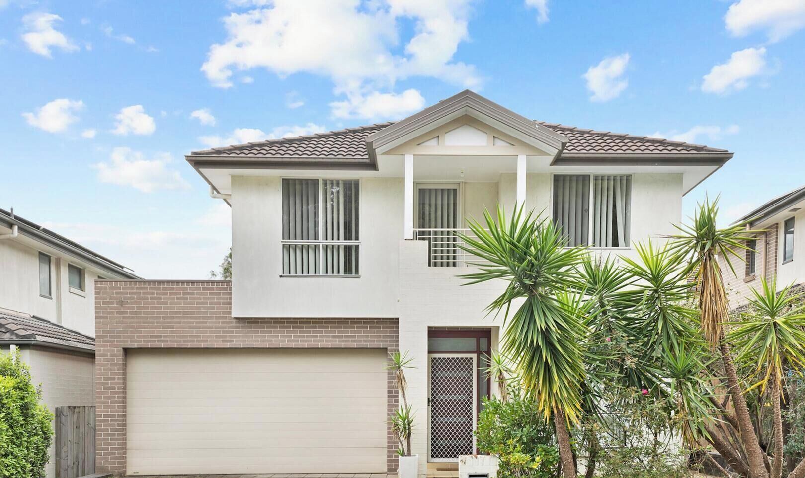 A two-story house with a garage, a balcony, and a small garden with palm trees.