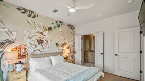 Ein Schlafzimmer mit einem großen Bett, einem Deckenventilator, einer Wand mit floralem Tapetenmuster und einer Tür zu einem Badezimmer.