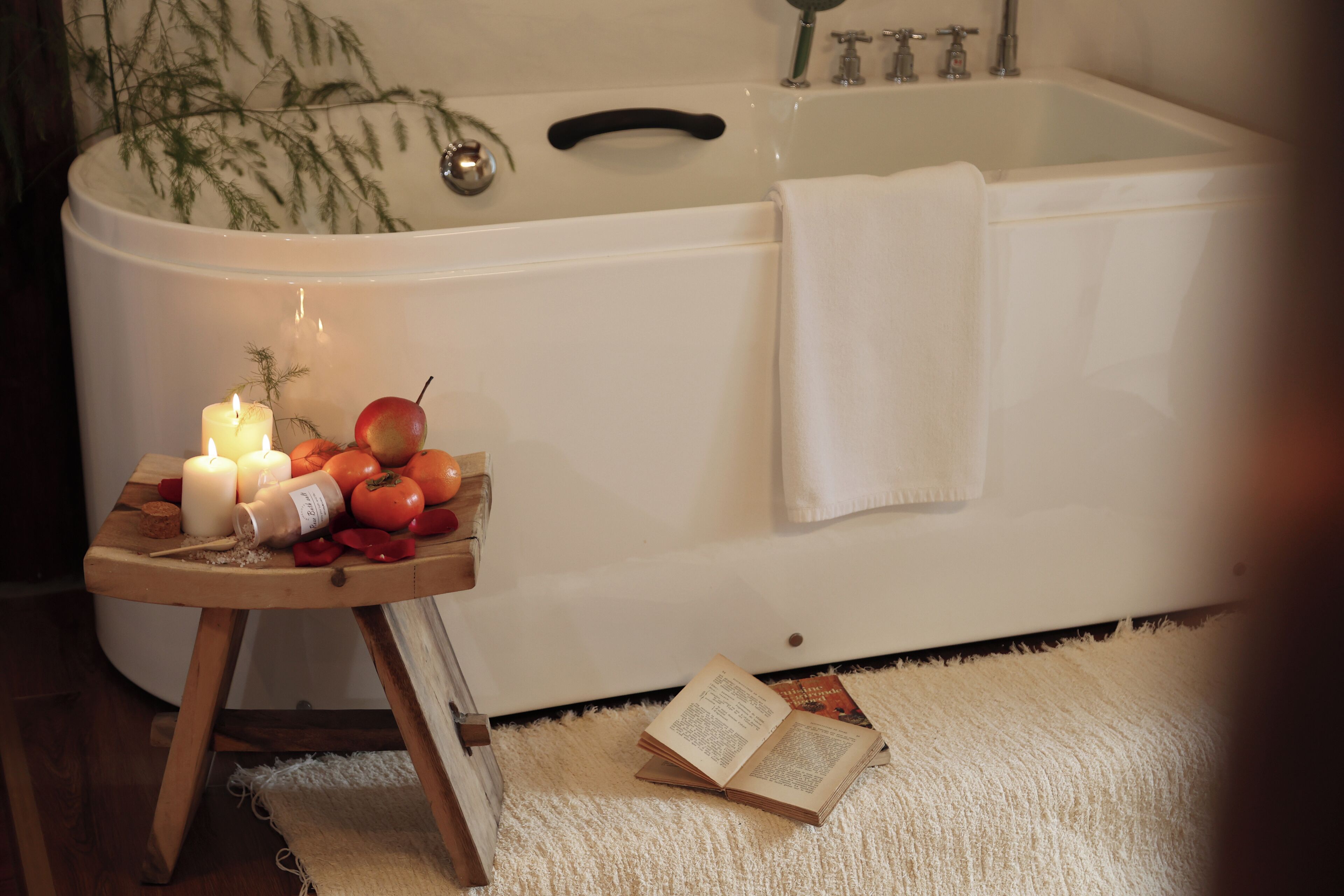 A bathtub with a towel, a wooden stool with candles and fruits, and an open book on a rug.