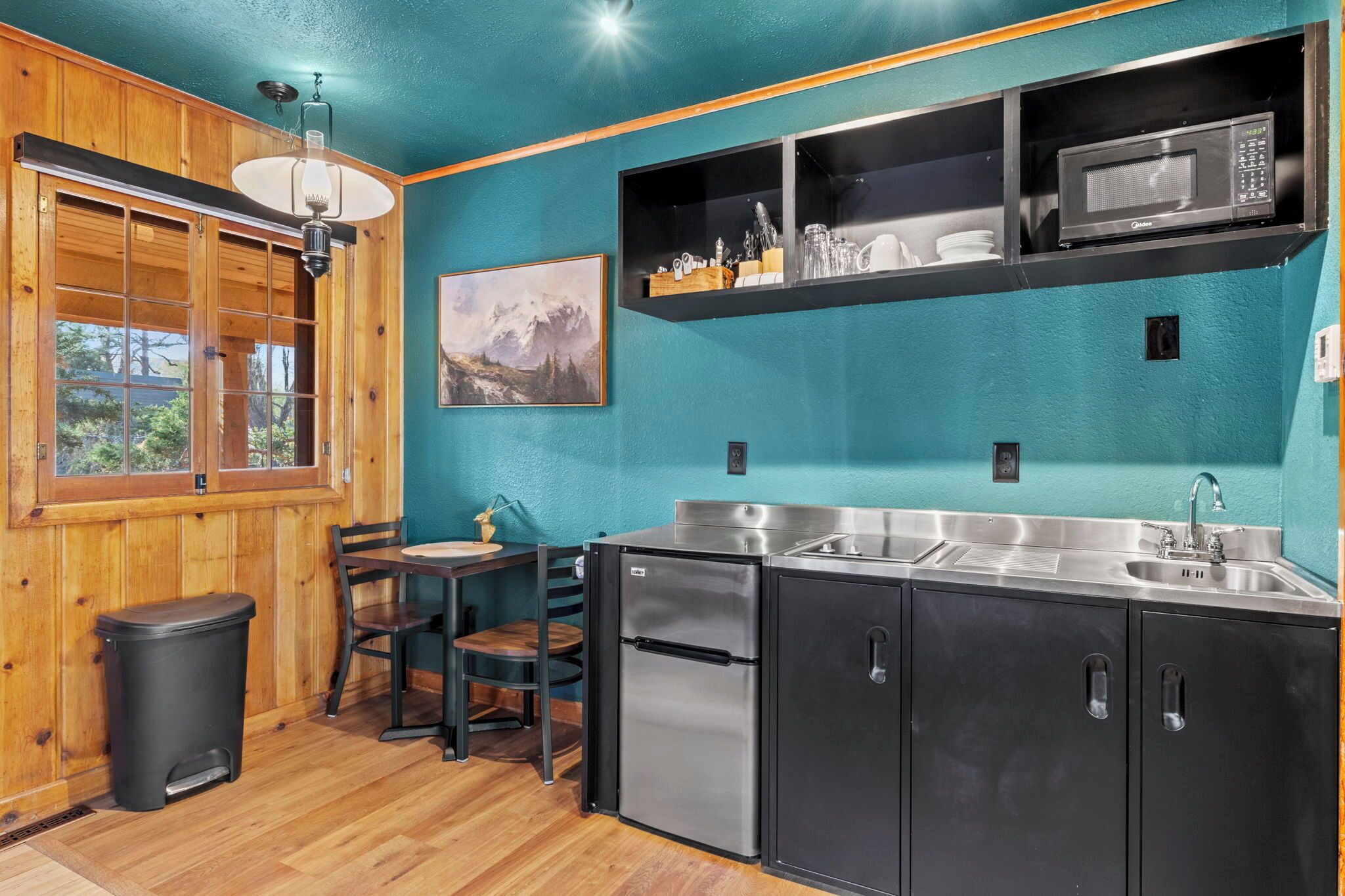 A compact kitchen with a stainless steel refrigerator, a microwave, and a sink. There is a wooden table with chairs and a trash bin.