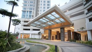 A modern hotel entrance with a covered walkway, a water feature, and a parking garage in the background. - Kenzi By SanLiving - 2BR - Kelapa Gading (Jakarta)