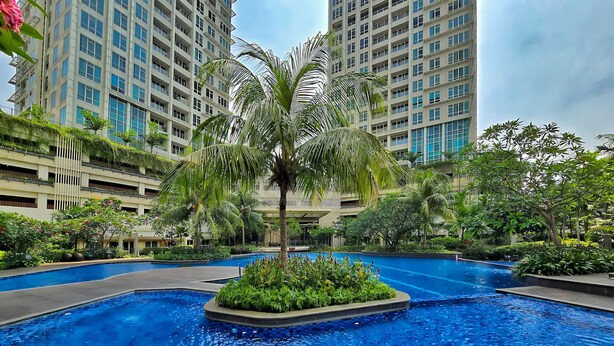 A swimming pool surrounded by lush greenery and tall buildings in the background. - Kenzi By SanLiving - 2BR - Kelapa Gading (Jakarta)