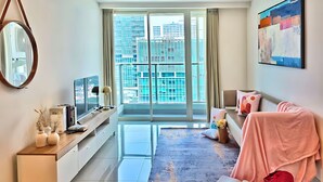 A modern bedroom with a large window, a flat-screen TV, a wooden console, a pink blanket, and a red basket. - Kenzi By SanLiving - 2BR - Kelapa Gading (Jakarta)