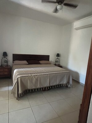 A bedroom with a bed, two bedside tables, a ceiling fan, and a wall-mounted air conditioner.