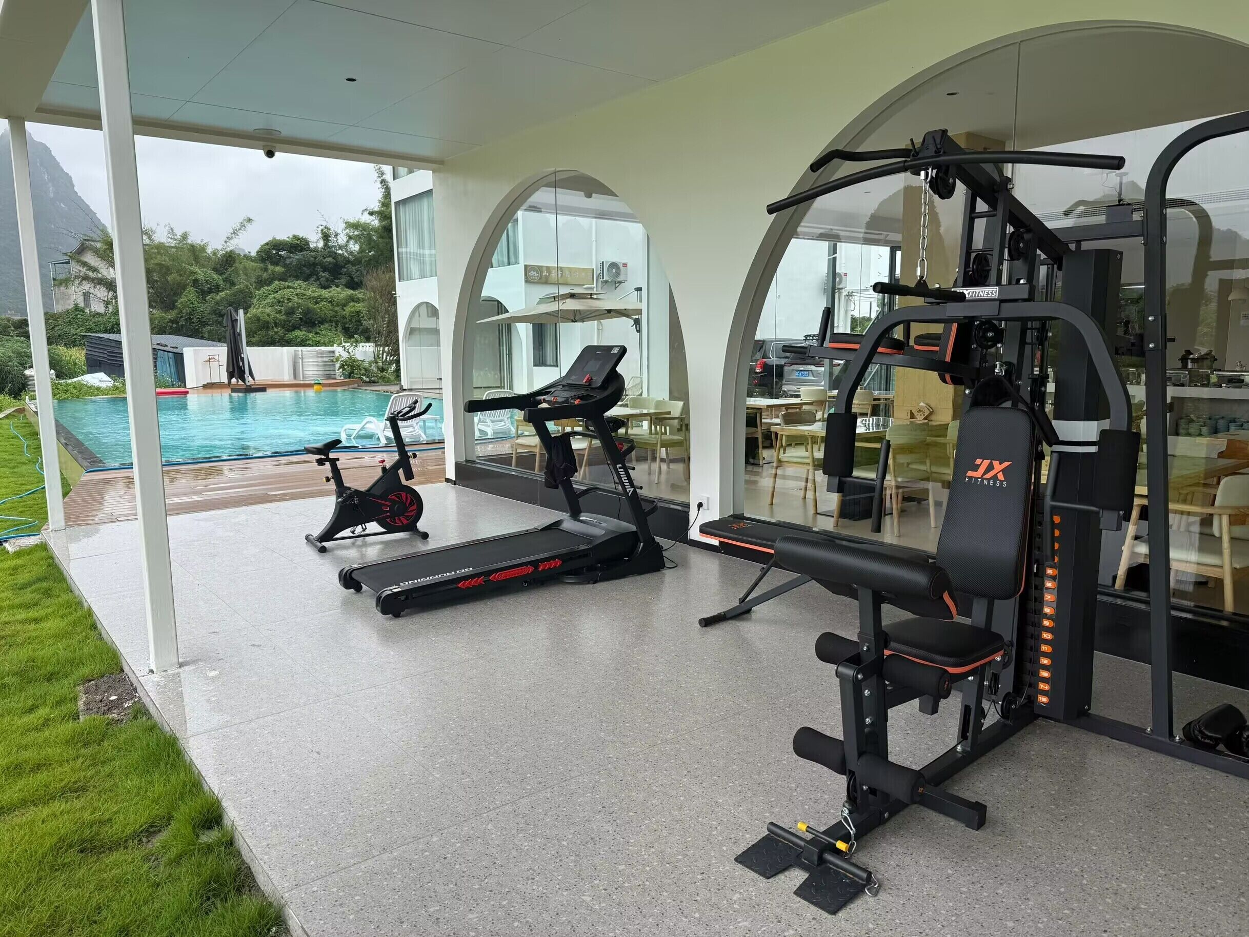 Een fitnessruimte met cardioapparatuur, een bank en een roeimachine. Door de glazen wanden is een zwembad zichtbaar.