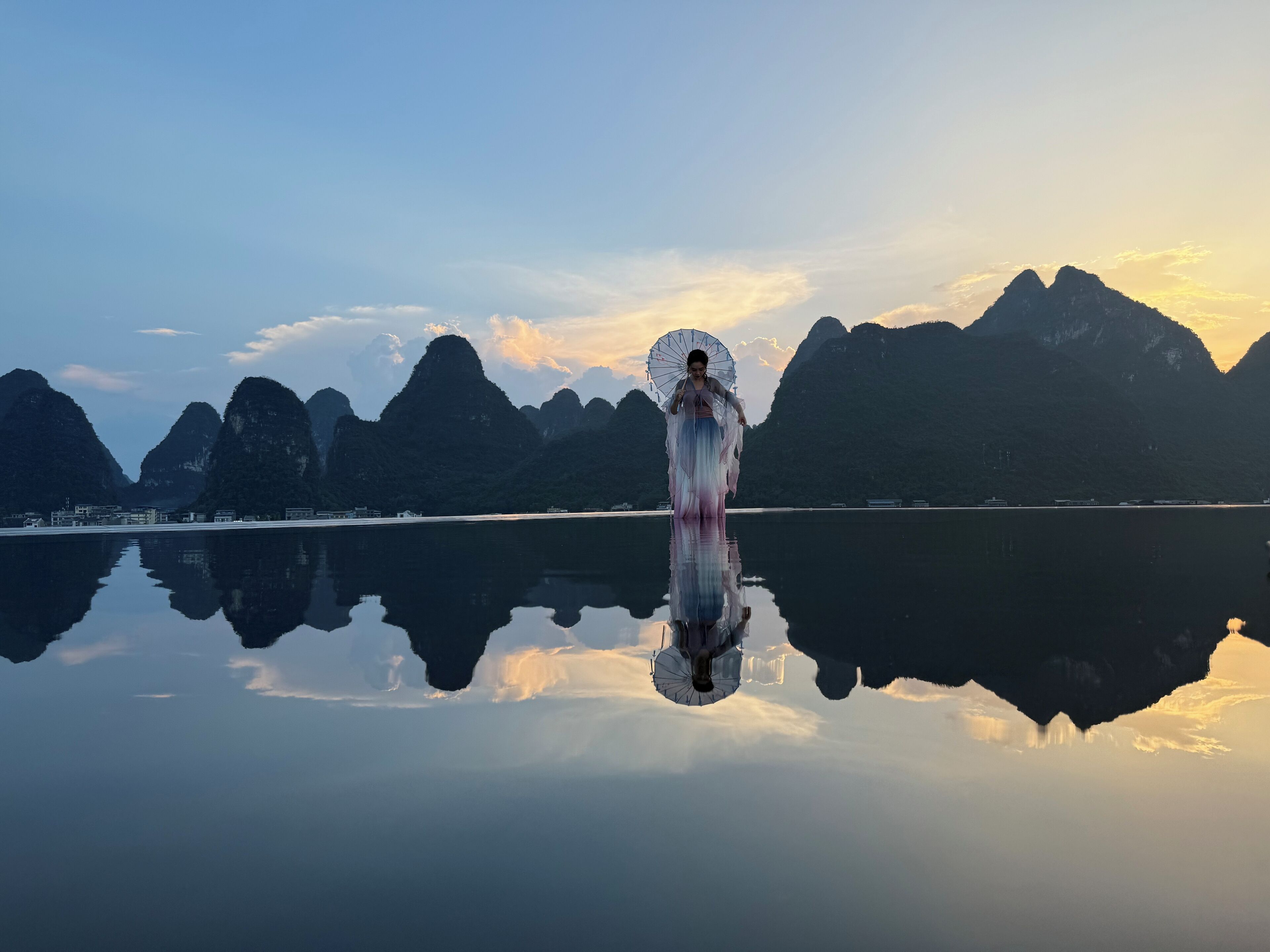 A person in a flowing dress standing by a calm lake with a mountain backdrop.