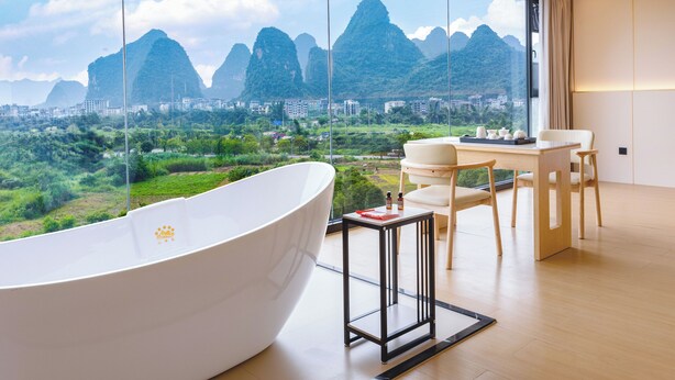 A modern bathroom with a large bathtub, a small table with toiletries, and a view of mountains and greenery. - Mountain Search Snow View Hotel (Guilin)