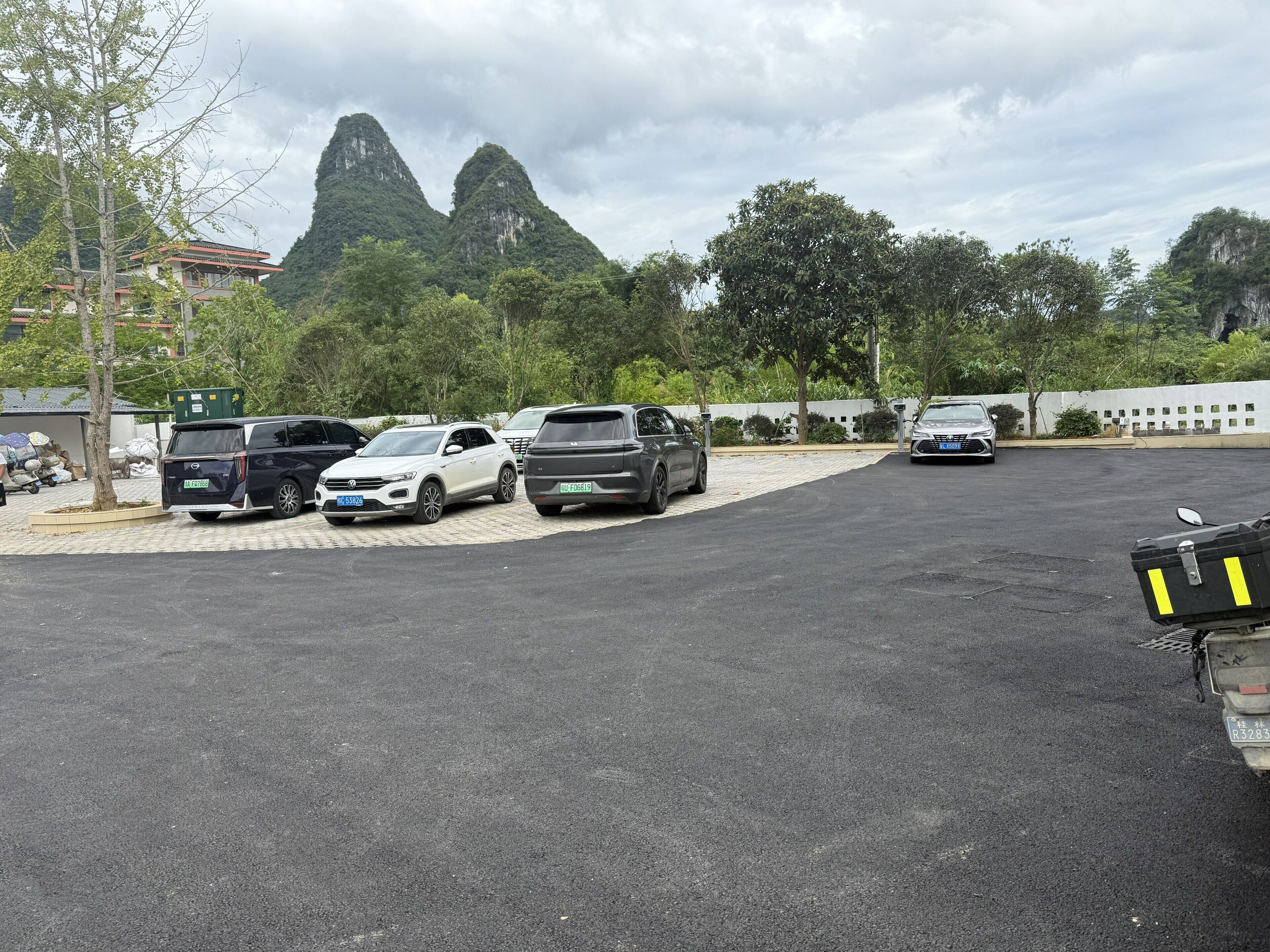 Um estacionamento com vários carros e uma motocicleta, cercado por árvores e um edifício com telhado tradicional.