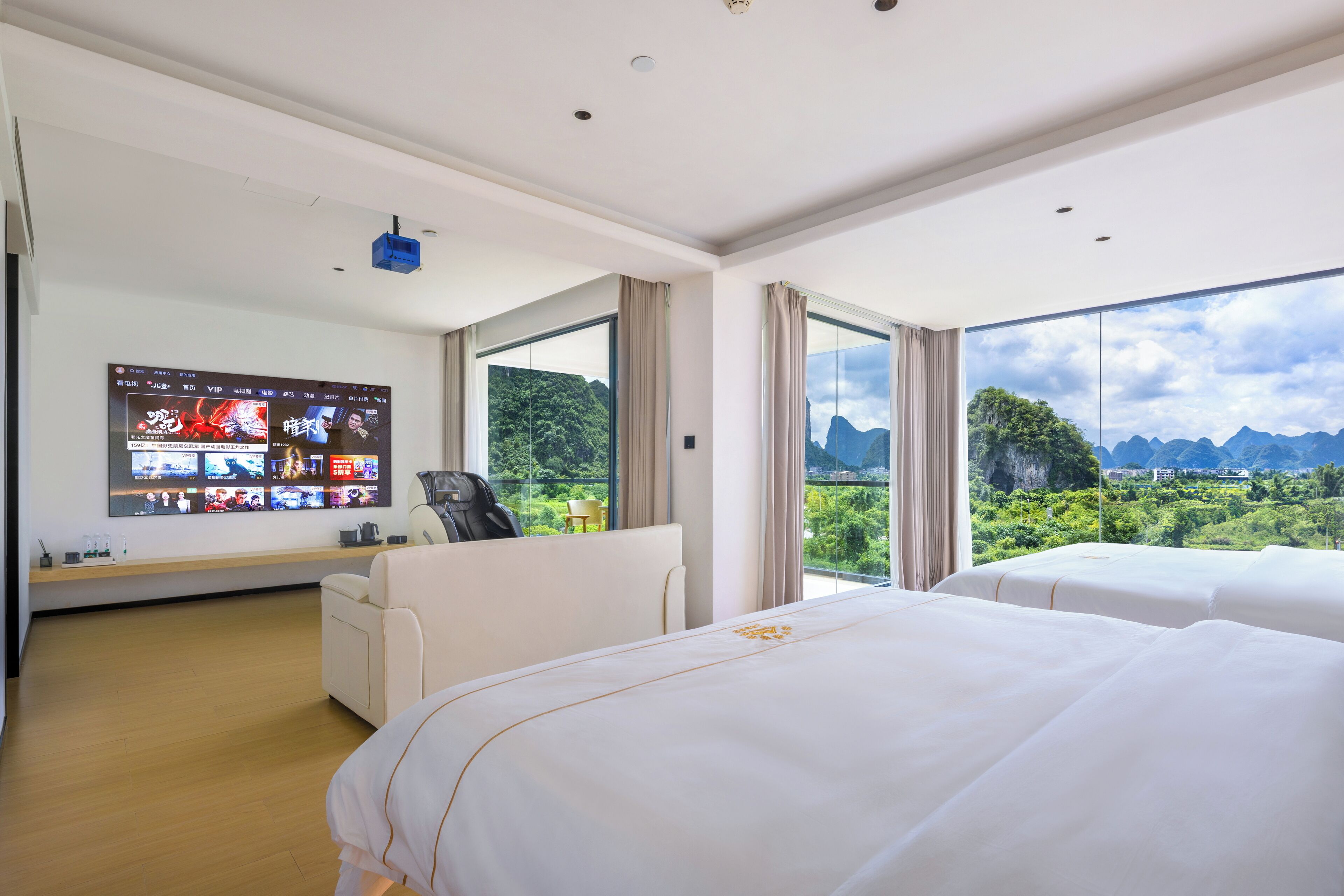 A hotel room with a large TV displaying multiple channels, a sofa, two beds, and a view of mountains and greenery.