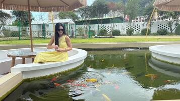 Una mujer sentada a una mesa redonda junto a un estanque con peces koi, rodeada de sombrillas y una valla.