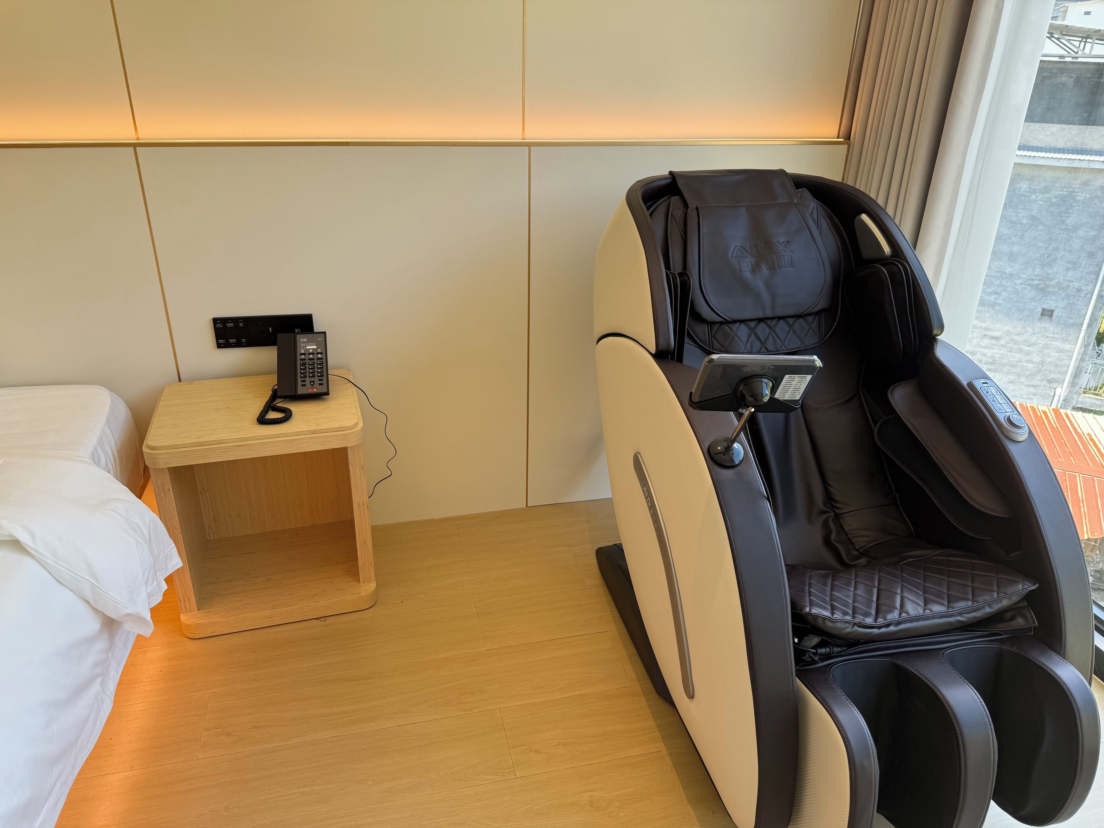 A hotel room with a massage chair, a bedside table with a phone, and a bed.