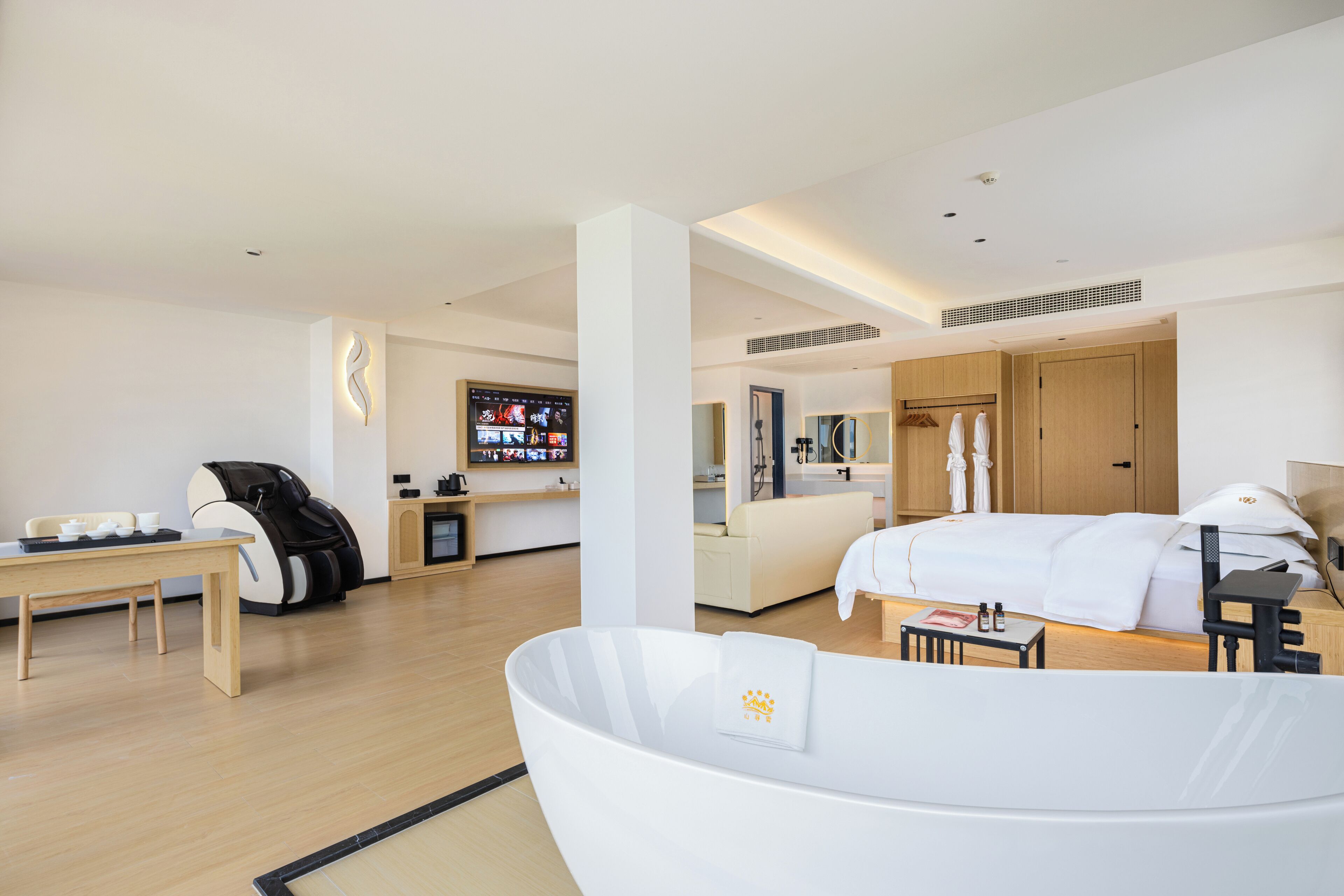 A modern hotel room with a large bed, a seating area with a massage chair, a small table with a coffee machine, and a TV mounted on the wall.