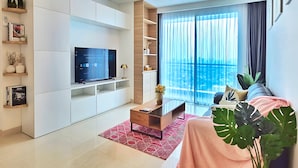 A modern living room with a flat-screen TV, built-in shelves, and a large window with curtains. - Amethyst By SanLiving - 3BR & 1 Maid Room - Central Park Mall (Jakarta)