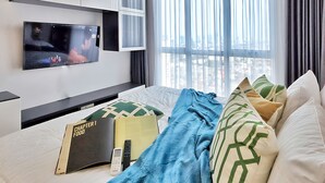 A modern hotel room with a large bed, a flat-screen TV mounted on the wall, a bedside table with a book and a remote control, and a window with sheer curtains. - Amethyst By SanLiving - 3BR & 1 Maid Room - Central Park Mall (Jakarta)