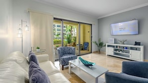 Ein modernes Wohnzimmer mit einer Couch, einem Sessel und einem Couchtisch. Ein Fernseher ist an der Wand befestigt und eine Glastür führt auf einen Balkon mit Pflanzen und Blick auf Gebäude.