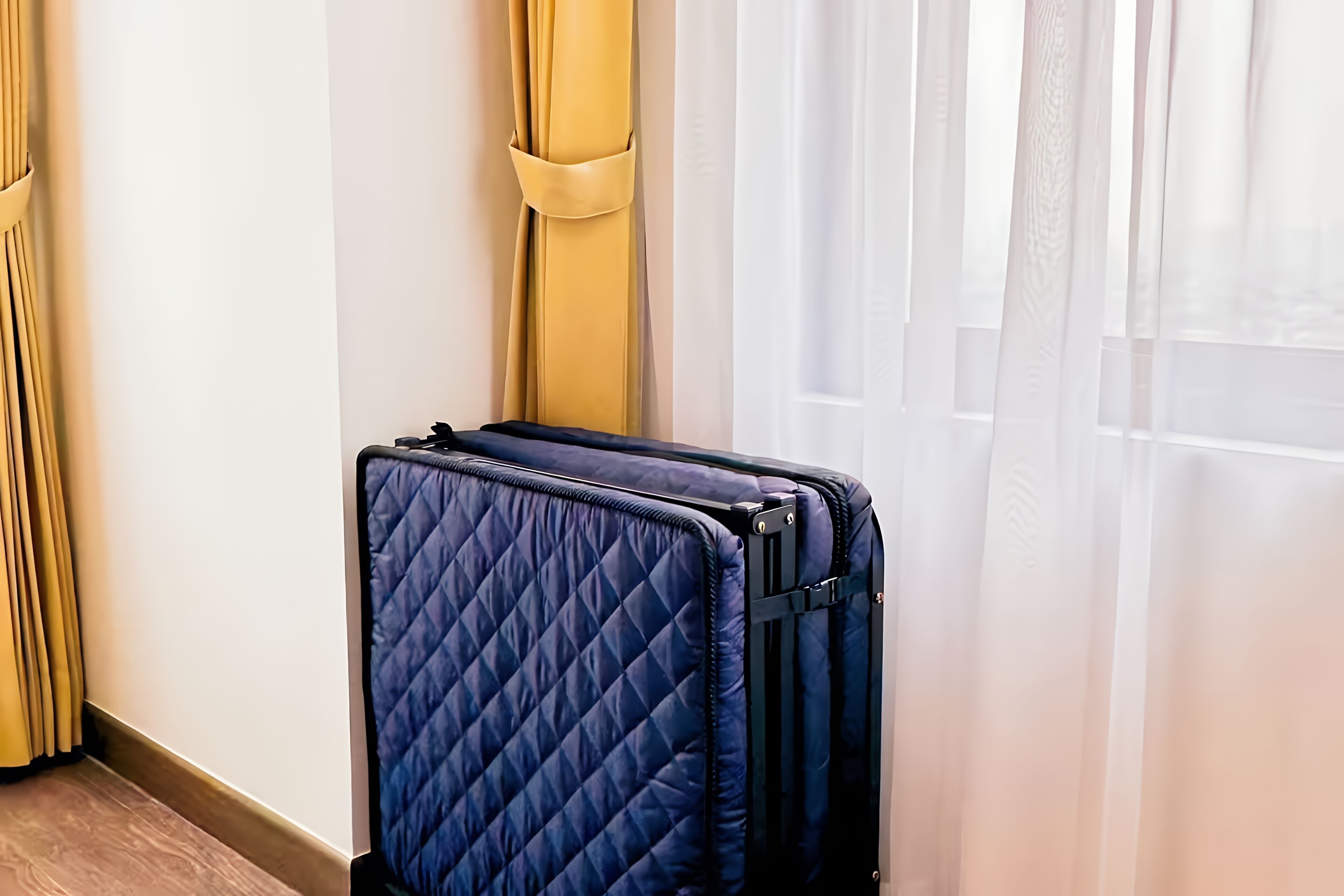 Une valise bleue matelassée, appuyée contre un mur avec une fenêtre et des rideaux.