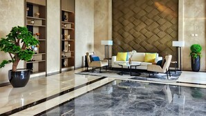 Un vestíbulo amplio con suelo de mármol, un gran árbol en maceta, un conjunto de sofás y un panel decorativo en la pared.
