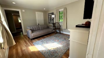 Ein Wohnzimmer mit grauem Sofa, einem gemusterten Teppich, Holzboden, einem Fenster mit Vorhängen und einer Tür zu einem anderen Raum.