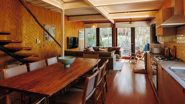 A modern kitchen with wooden cabinetry, a dining area with a wooden table and chairs, and a staircase with wooden steps.