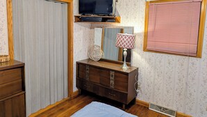 Une chambre avec une commode en bois, un téléviseur posé sur un meuble en bois, une lampe de chevet, une fenêtre avec des stores et un radiateur.