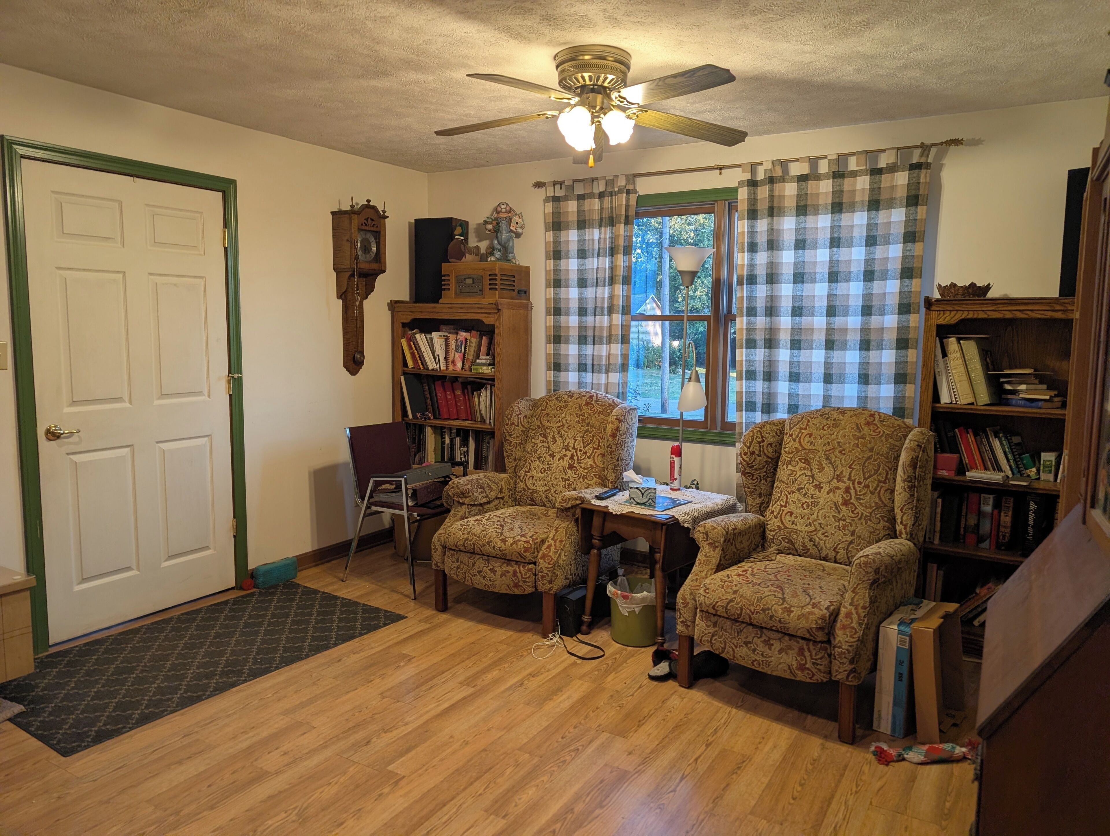A cozy living room with two patterned armchairs, a wooden bookshelf, a ceiling fan, and a window with plaid curtains.