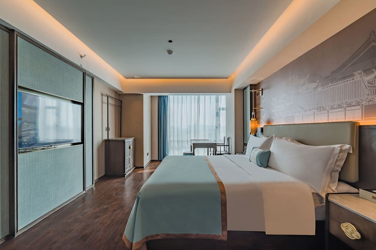A modern hotel room with a large bed, a desk, and a view of a cityscape through large windows.