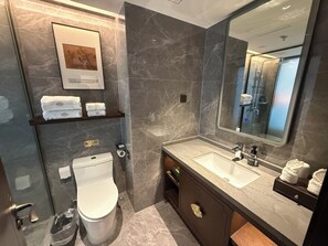 A modern bathroom with a marble wall, a large mirror, a sink, and a toilet. There are towels on a shelf and a trash bin. - Till Bright Hotel  (Foshan)