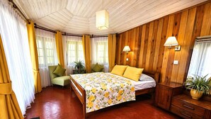 A room with a wooden ceiling, a bed with yellow bedding, a green chair, a bedside table with a lamp, and a wooden cabinet with a plant.