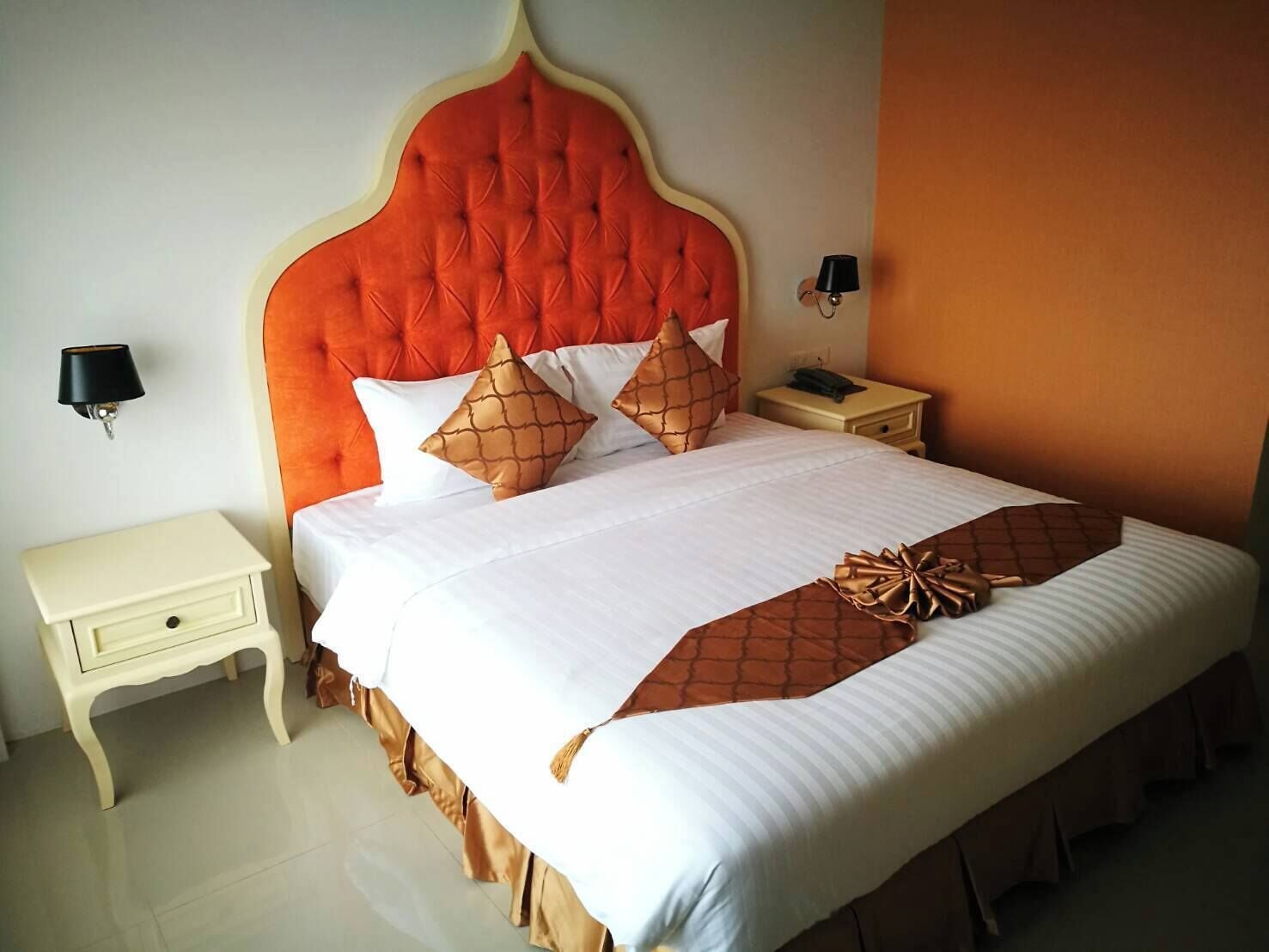 Una cama bien hecha con un cabecero capitonado, ropa de cama blanca y almohadas decorativas. Hay mesitas de noche con lámparas y un aparador con un teléfono.