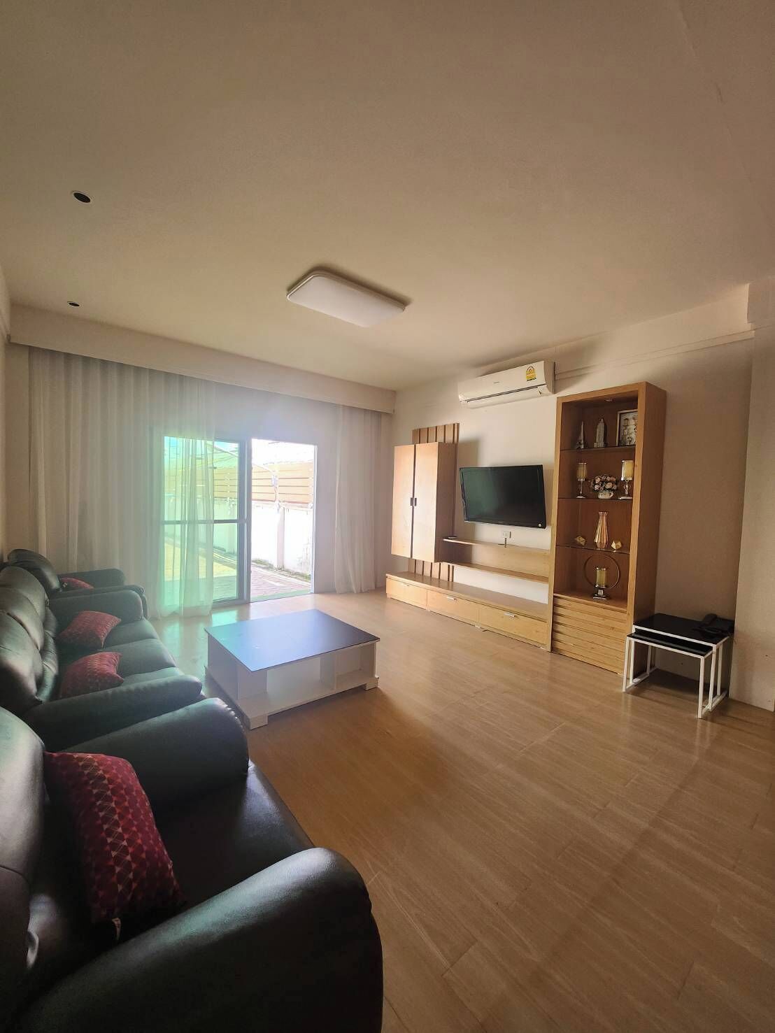 A modern living room with a leather sofa, a flat-screen TV, wooden cabinets, and a sliding glass door leading to an outdoor area.