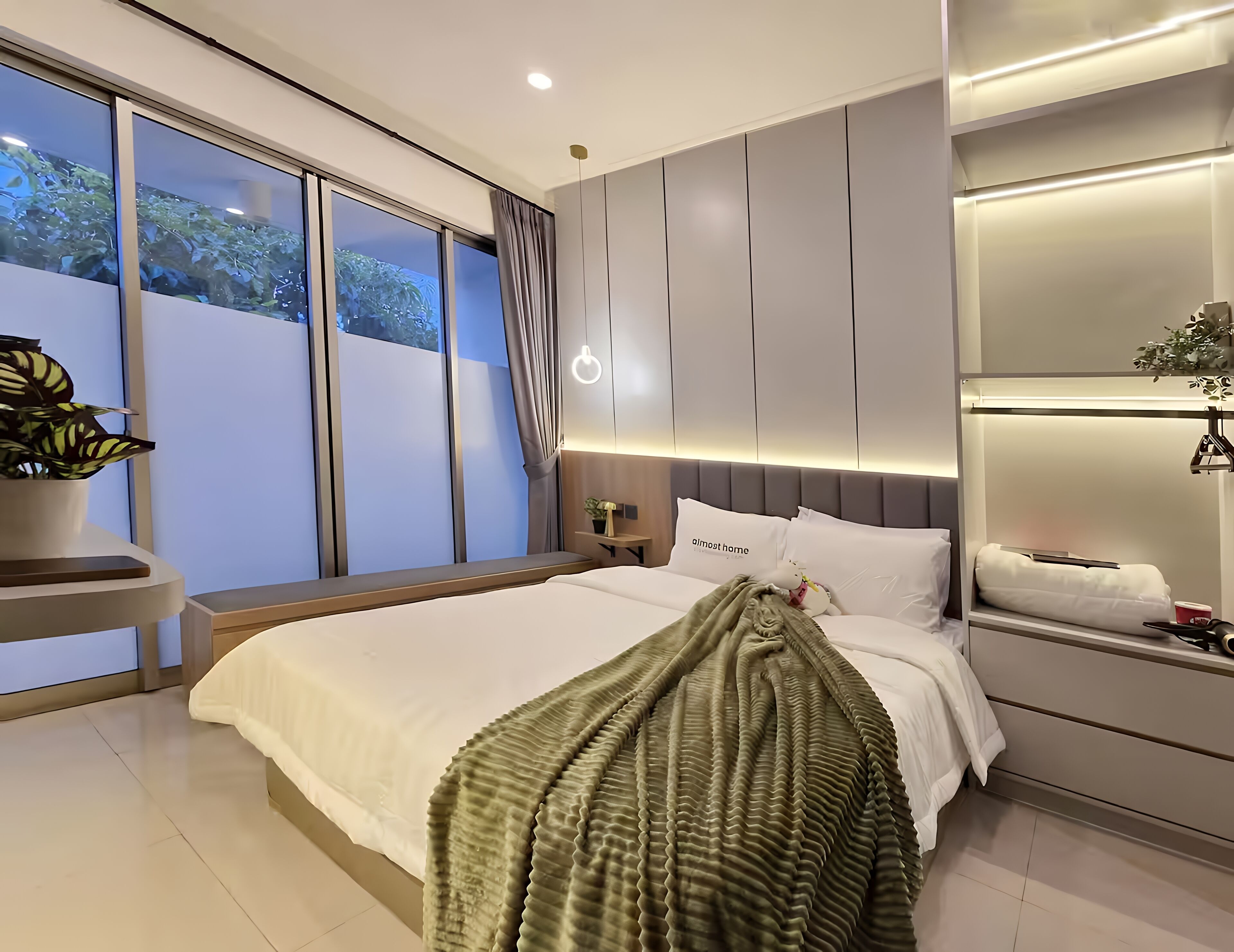 A modern bedroom with a large bed, a bedside table, a nightstand, a potted plant, and a view of greenery outside.