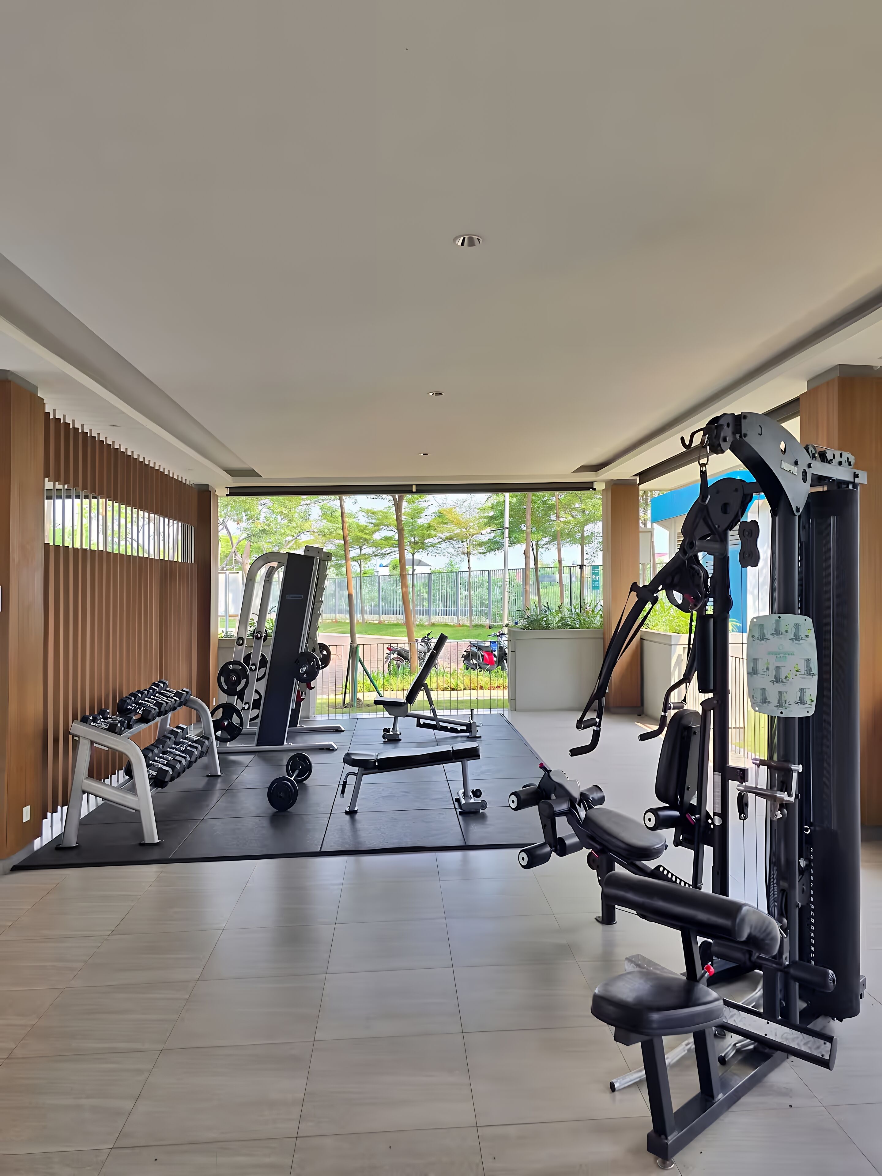 A gym with various exercise equipment, a bench, dumbbells, and a view of a park through large windows.