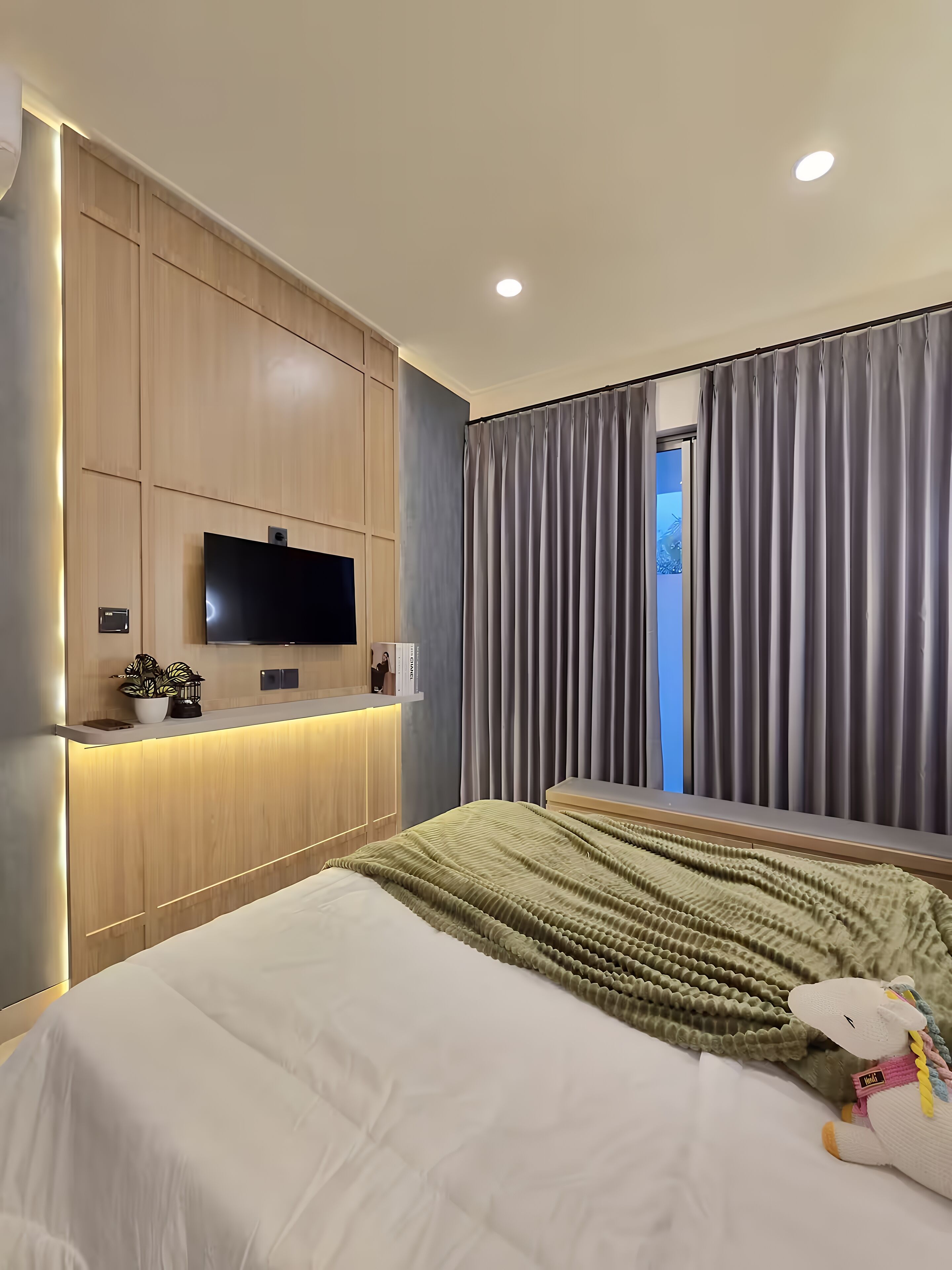Una habitación de hotel moderna con una cama grande, un televisor de pantalla plana montado en un panel de madera, una mesita de noche con una lámpara y vistas al exterior a través de una ventana con cortinas grises.
