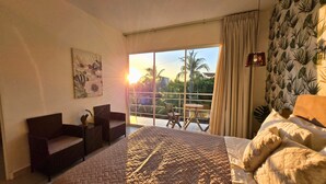 Une chambre dotée d’un grand lit, d’un canapé, d’une petite table et d’un balcon offrant une vue sur les palmiers et le coucher de soleil.