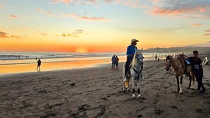 Une scène de plage avec des chevaux et des cavaliers, un coucher de soleil, et des personnes en arrière-plan.