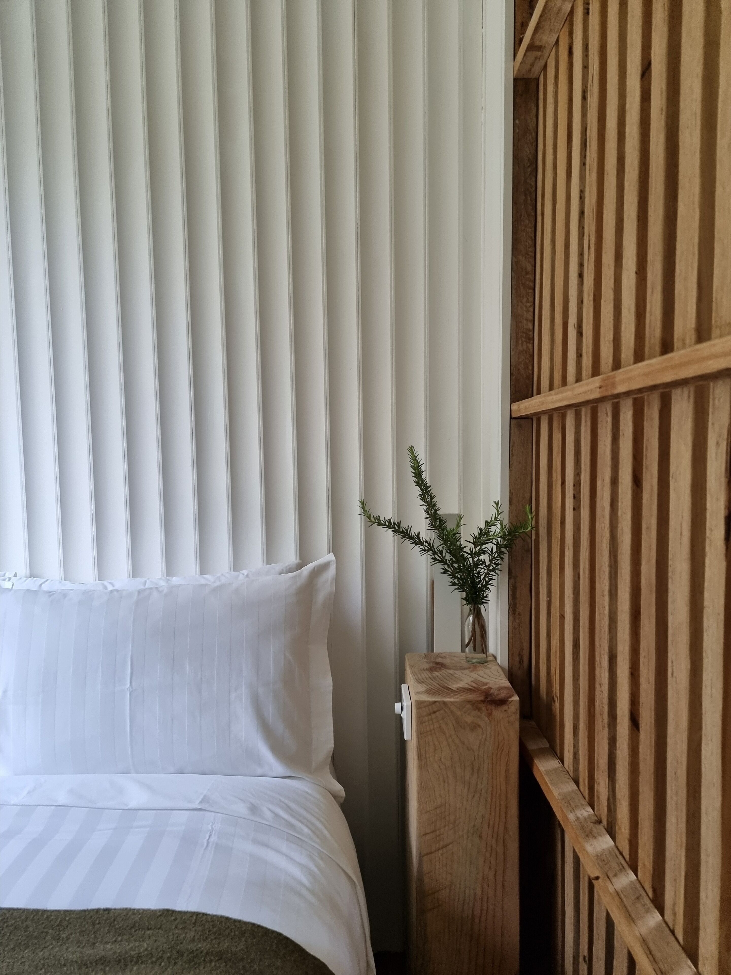 A bedroom with a bed, white curtains, a wooden headboard, and a small plant on a wooden nightstand.