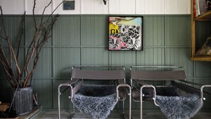 A room with two metal-framed chairs with fur throw pillows, a green wall, a framed abstract painting, and a wooden bookshelf.