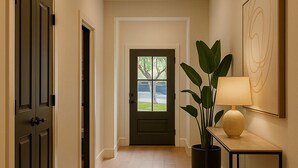 Un pasillo con suelo de madera, una puerta oscura, una puerta con paneles de vidrio, una planta en maceta, una mesa con lámpara y una obra de arte enmarcada en la pared.