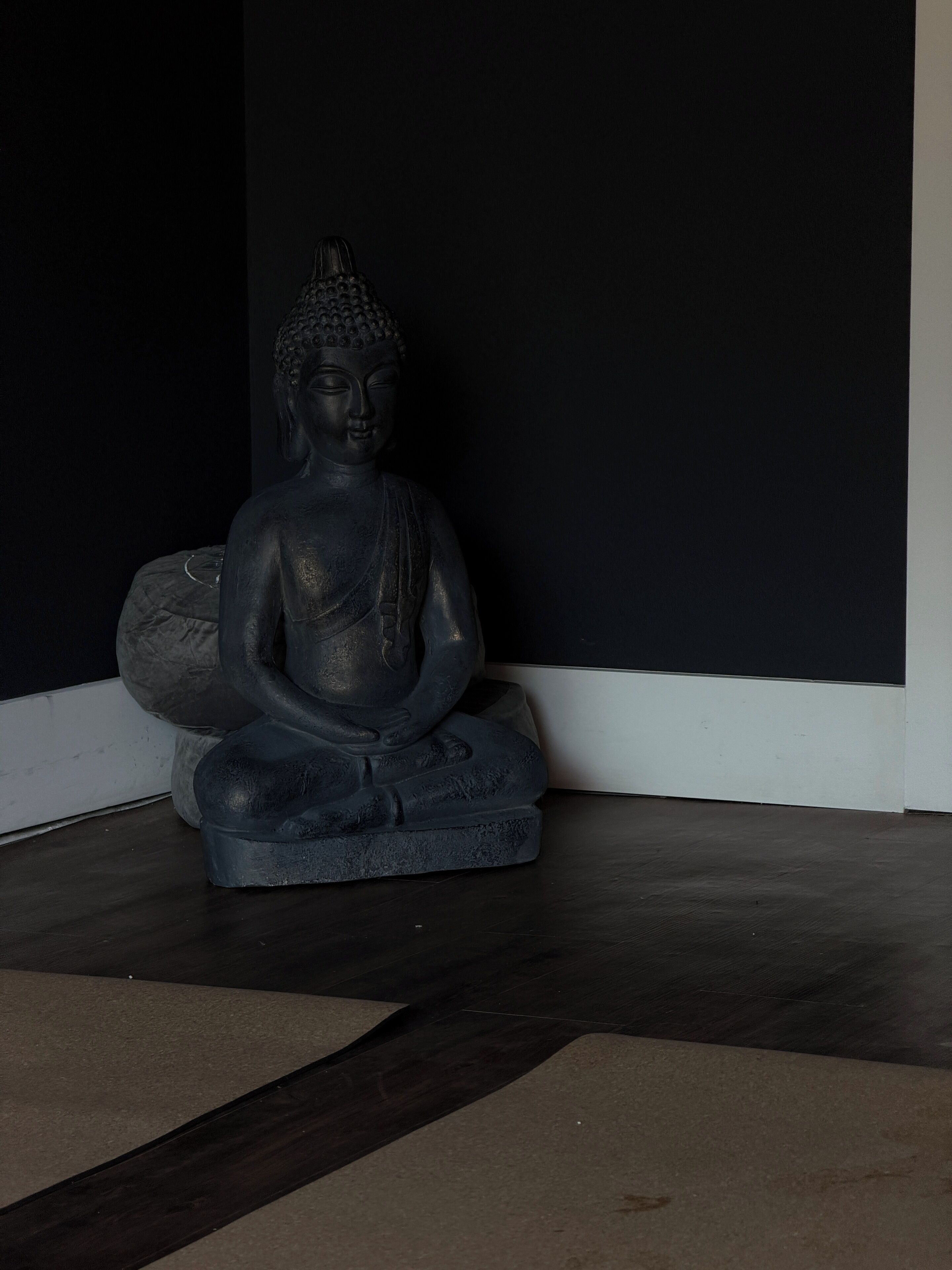 Una estatua oscura de Buda en un rincón de una habitación con suelo oscuro y pared blanca.