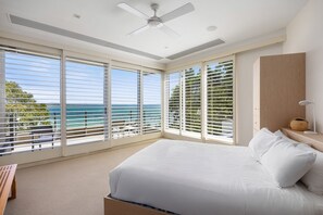 A bedroom with a large bed, a ceiling fan, and a view of the ocean through floor-to-ceiling windows.