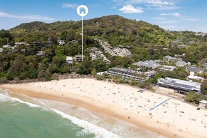 A coastal area with a beach, a hotel with multiple floors, and a hill with dense vegetation in the background.