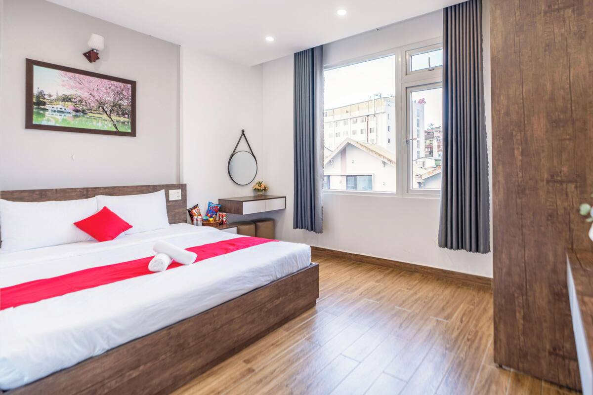 A hotel room with a bed, a desk, a window with curtains, a wooden floor, and a painting on the wall.