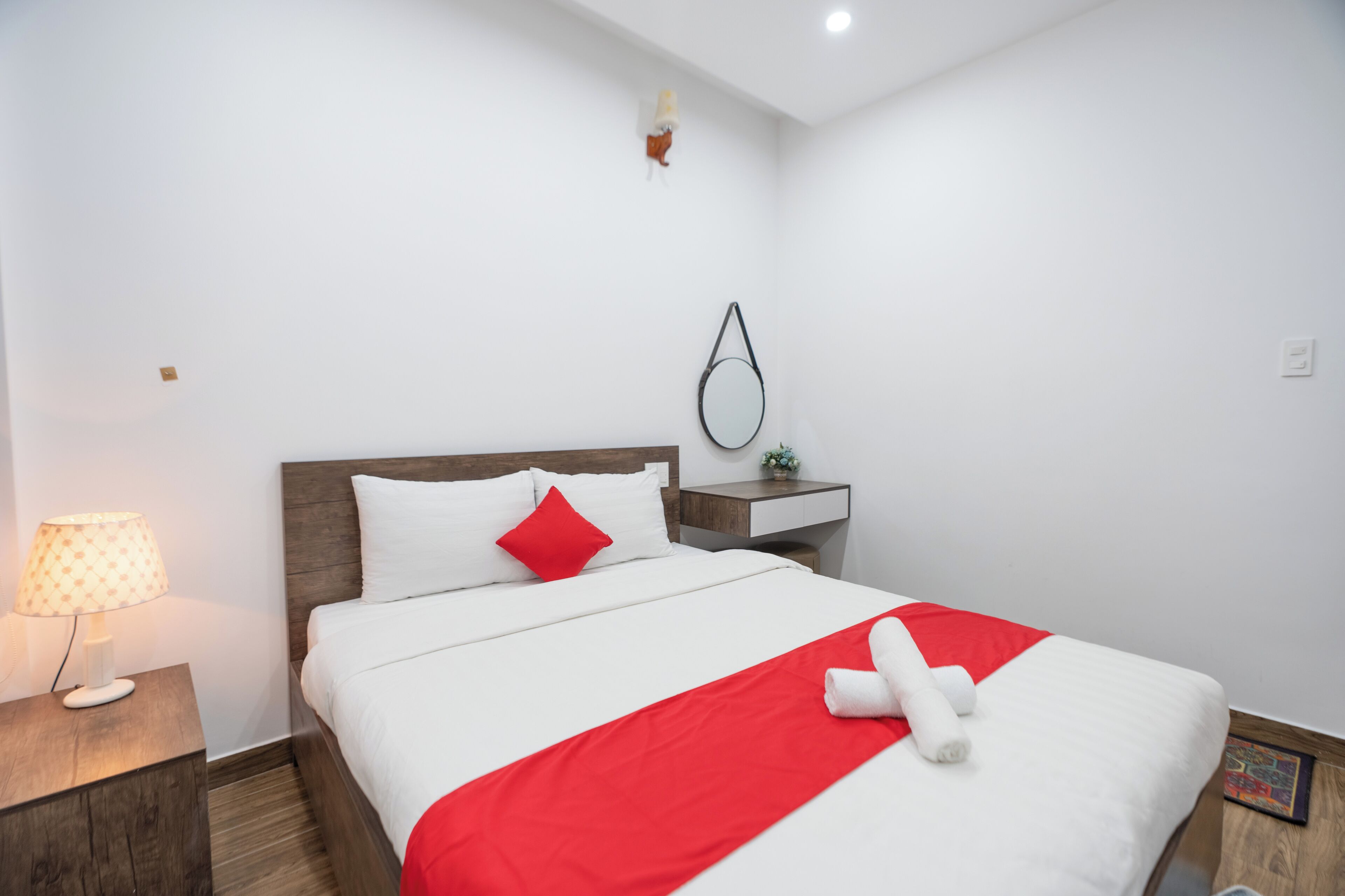 Cama bem arrumada com roupa de cama branca e vermelha, abajur, mesa de cabeceira pequena e espelho fixado na parede.
