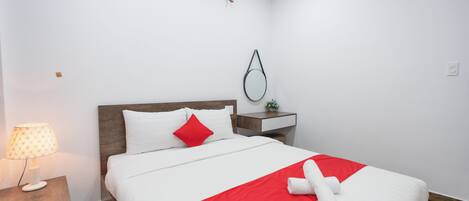 Cama bem arrumada com roupa de cama branca e vermelha, abajur, mesa de cabeceira pequena e espelho fixado na parede.