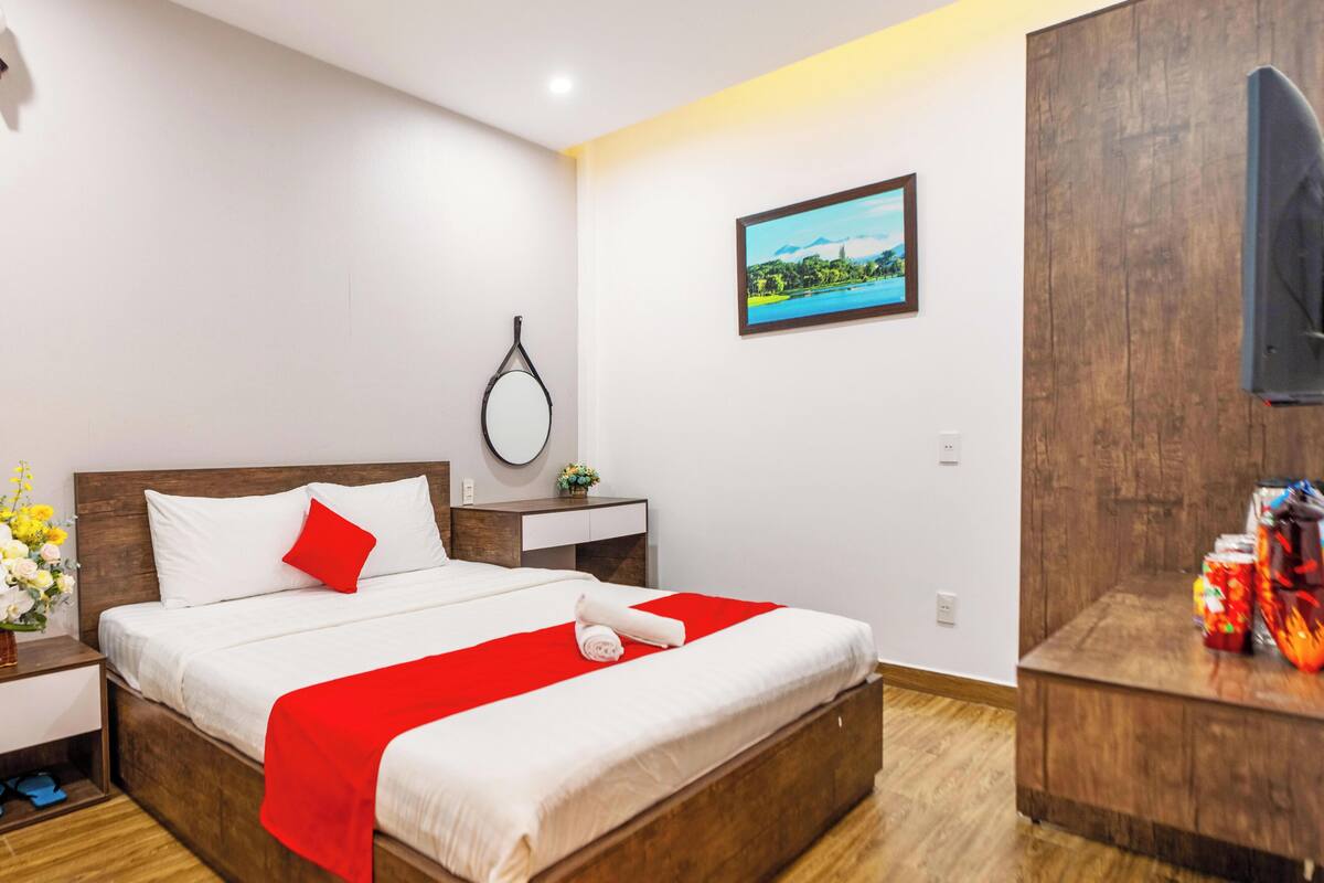 A hotel room with a wooden bed, a red throw, a small bedside table with a vase of flowers, a wall-mounted TV, and a framed picture.