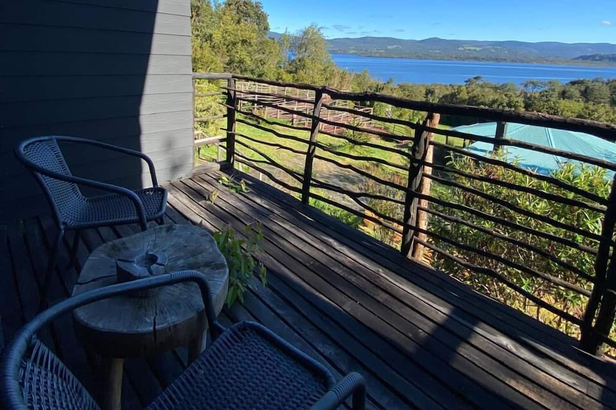 A wooden deck with two wicker chairs and a small round table, overlooking a lake with a distant shoreline.