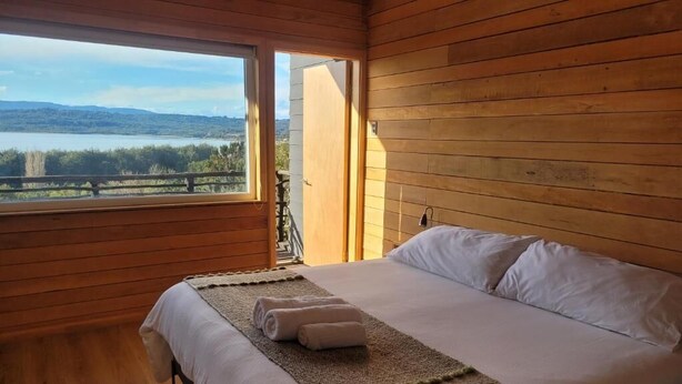 A wooden cabin bedroom with a bed, a view of a lake, and a balcony. - Huillín Lodge (Chonchi)